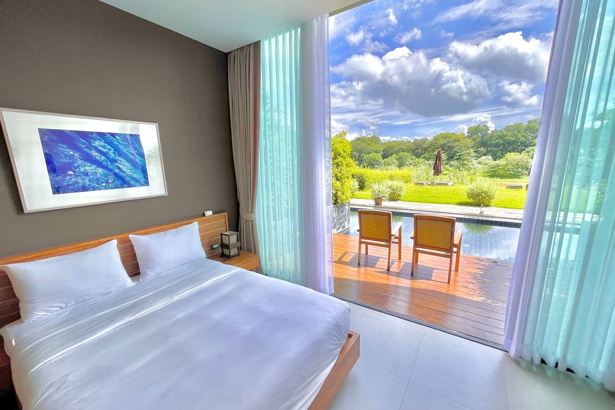 Quarto de hotel com uma cama grande, um quadro na parede e vista para a piscina e jardim pela janela.