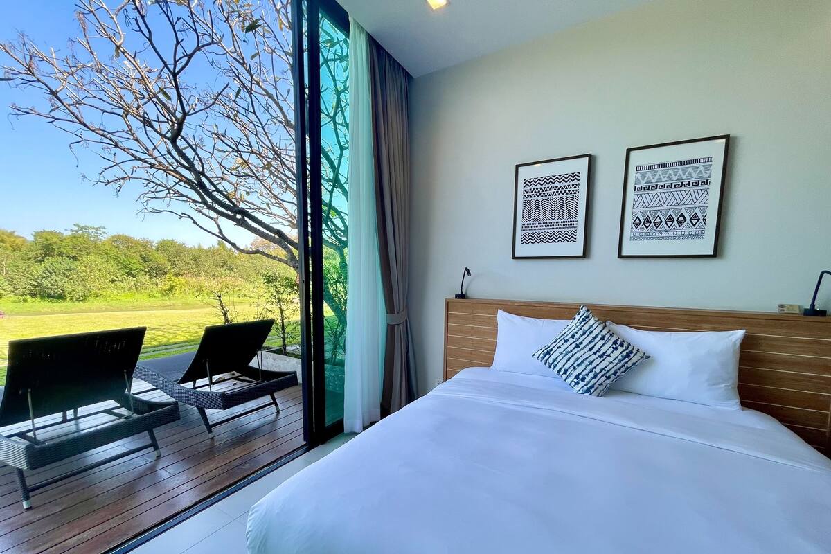 Quarto com cama grande, duas fotos emolduradas na parede e vista para uma área gramada através de uma porta de vidro.