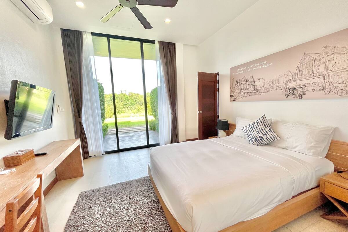 Quarto com cama, televisão, ventoinha no teto, escrivaninha e uma janela grande com cortinas.