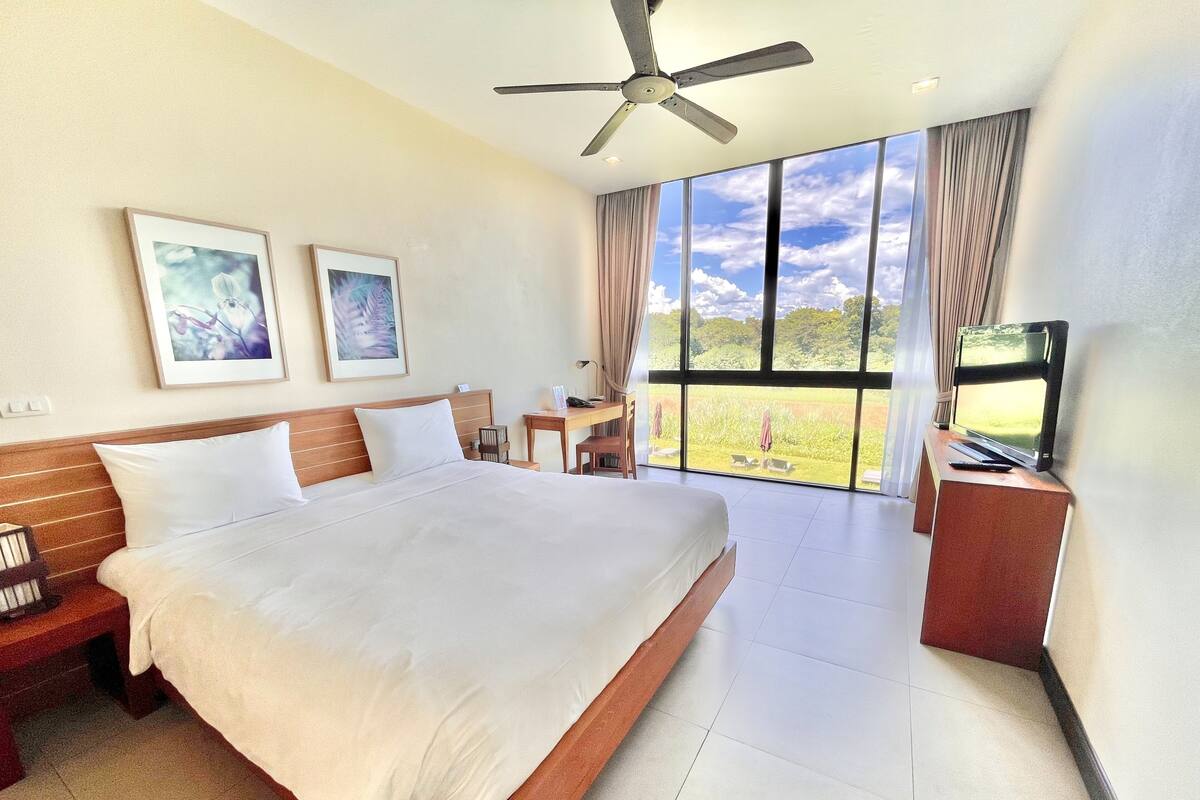 Quarto espaçoso com uma cama grande, uma escrivaninha, uma televisão e vista para uma paisagem verde através de grandes janelas.