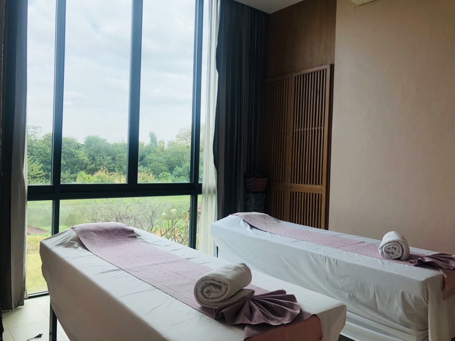 A room with two massage tables, large windows with a view of greenery, and a wooden wardrobe.