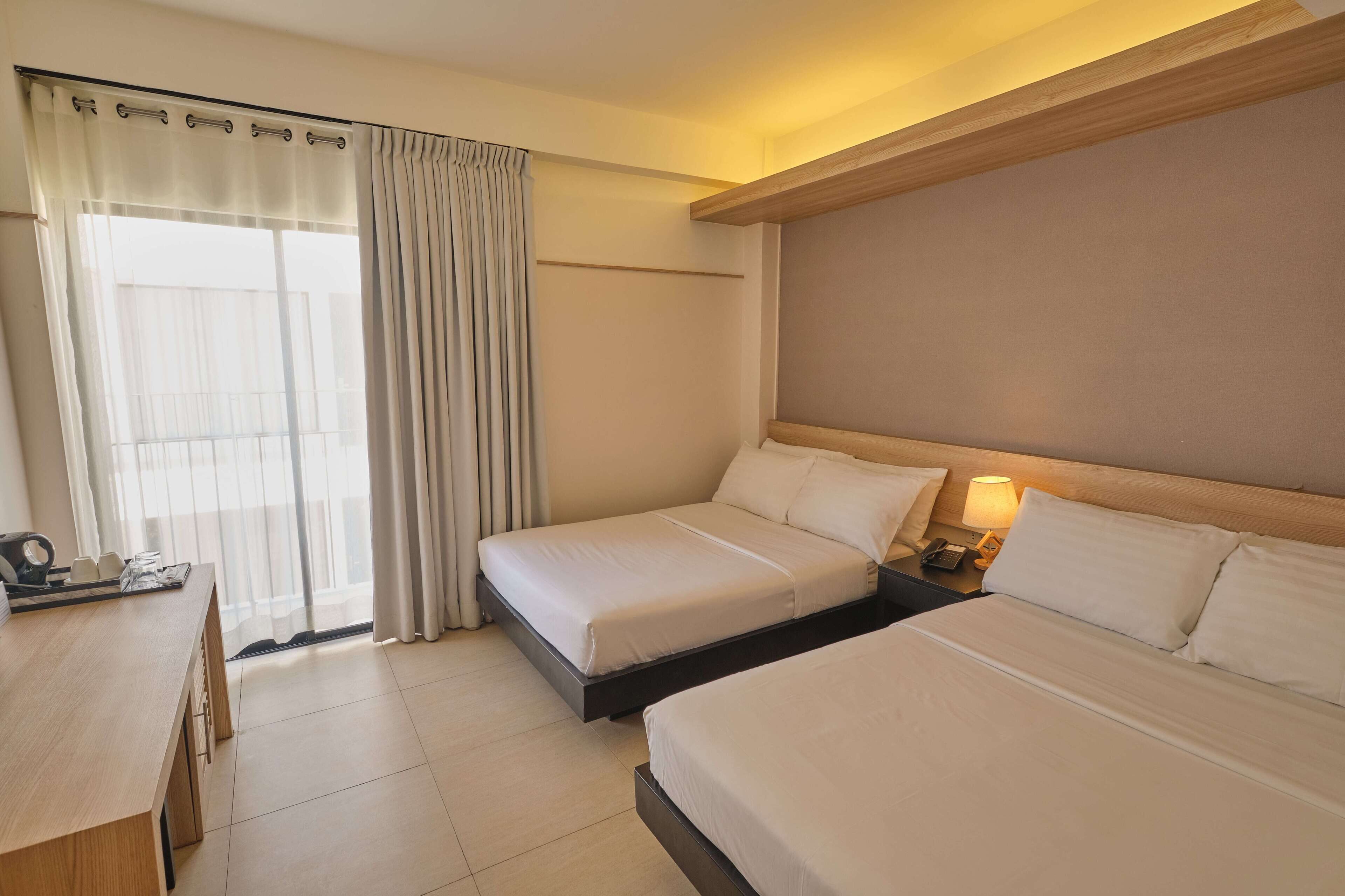 A hotel room with two beds, a desk, a lamp, and a window with curtains.