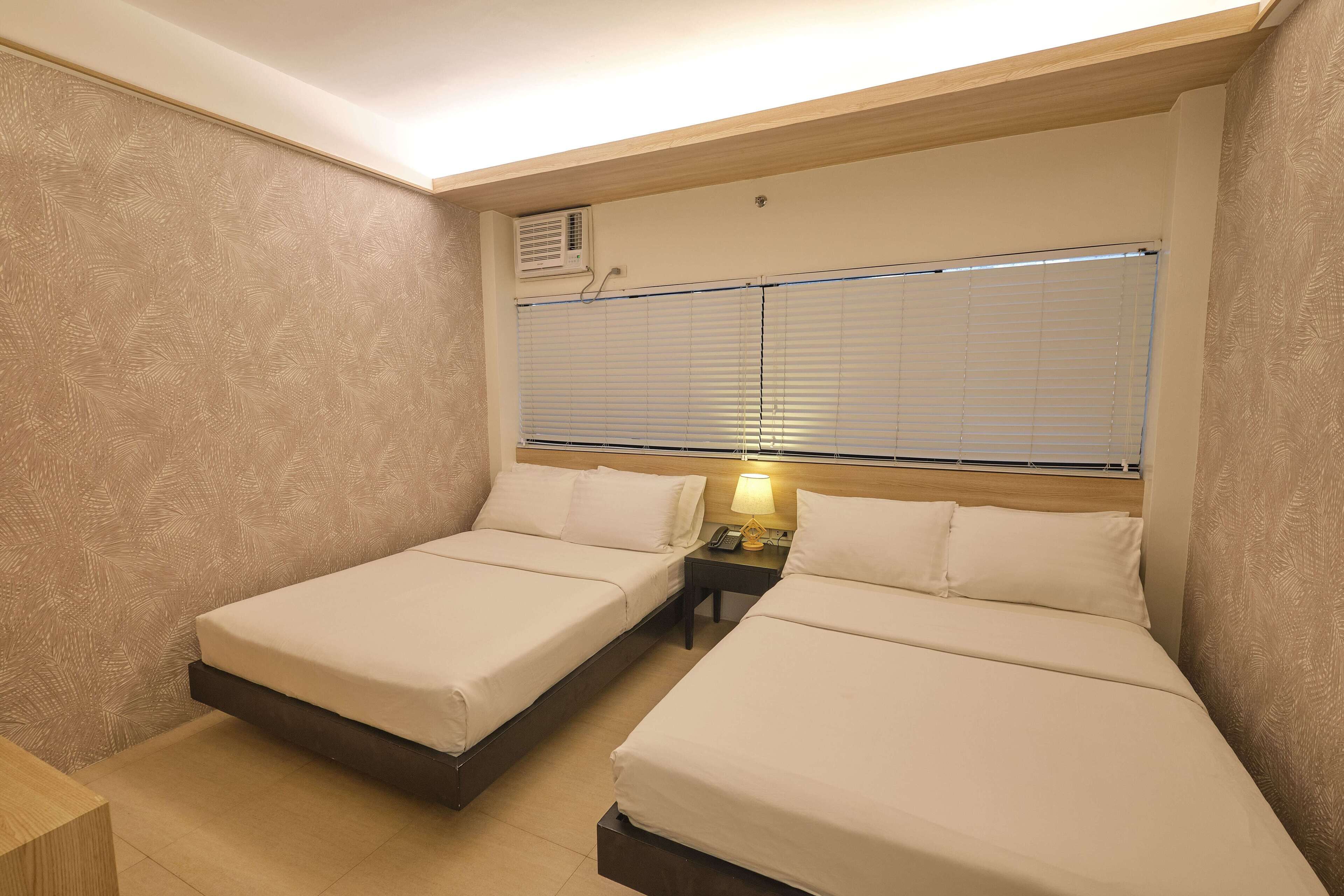 Habitación de hotel con dos camas individuales, una mesita de noche con lámpara, una ventana con persianas y un equipo de aire acondicionado en la pared.