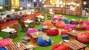 Un espacio al aire libre con mesas y sillas de madera, coloridos puffs y una zona de bar con una persona atendiendo.