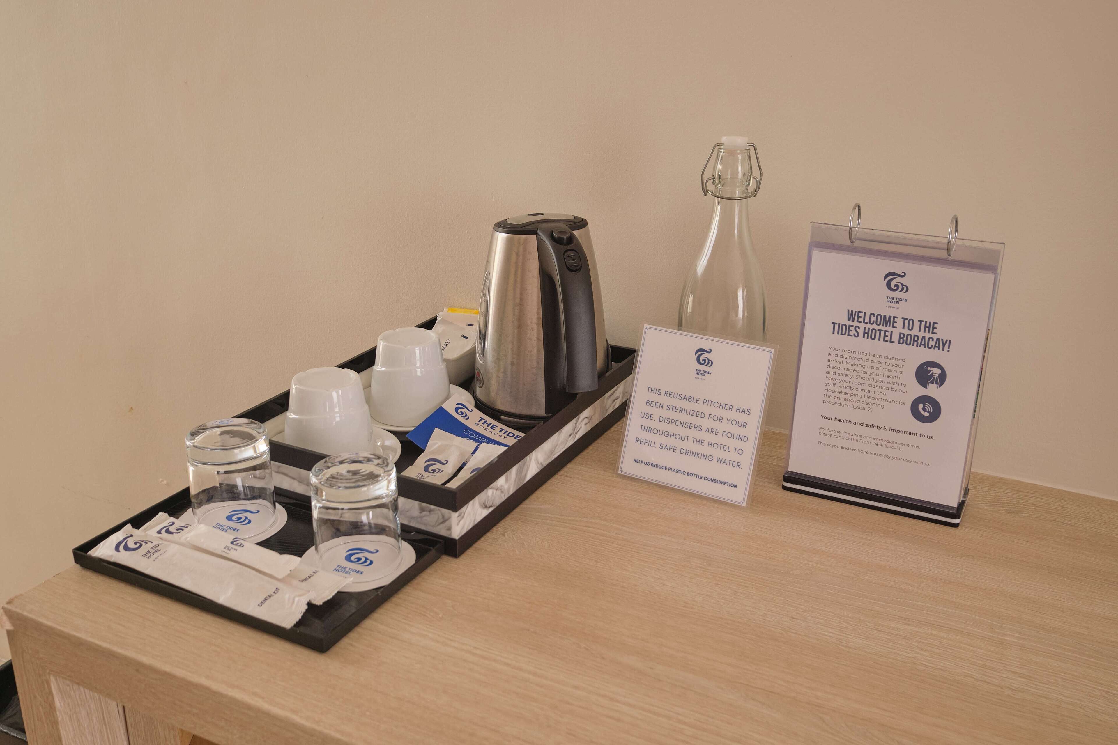 A hotel room's breakfast tray with a coffee maker, teapot, pitcher, cups, and condiments. A welcome sign and a refillable water bottle are also visible.
