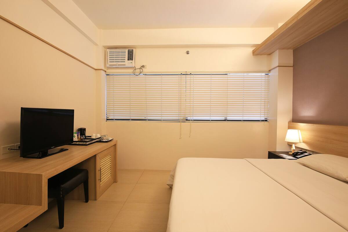 A hotel room with a bed, a desk with a television, a lamp, and a window with blinds.