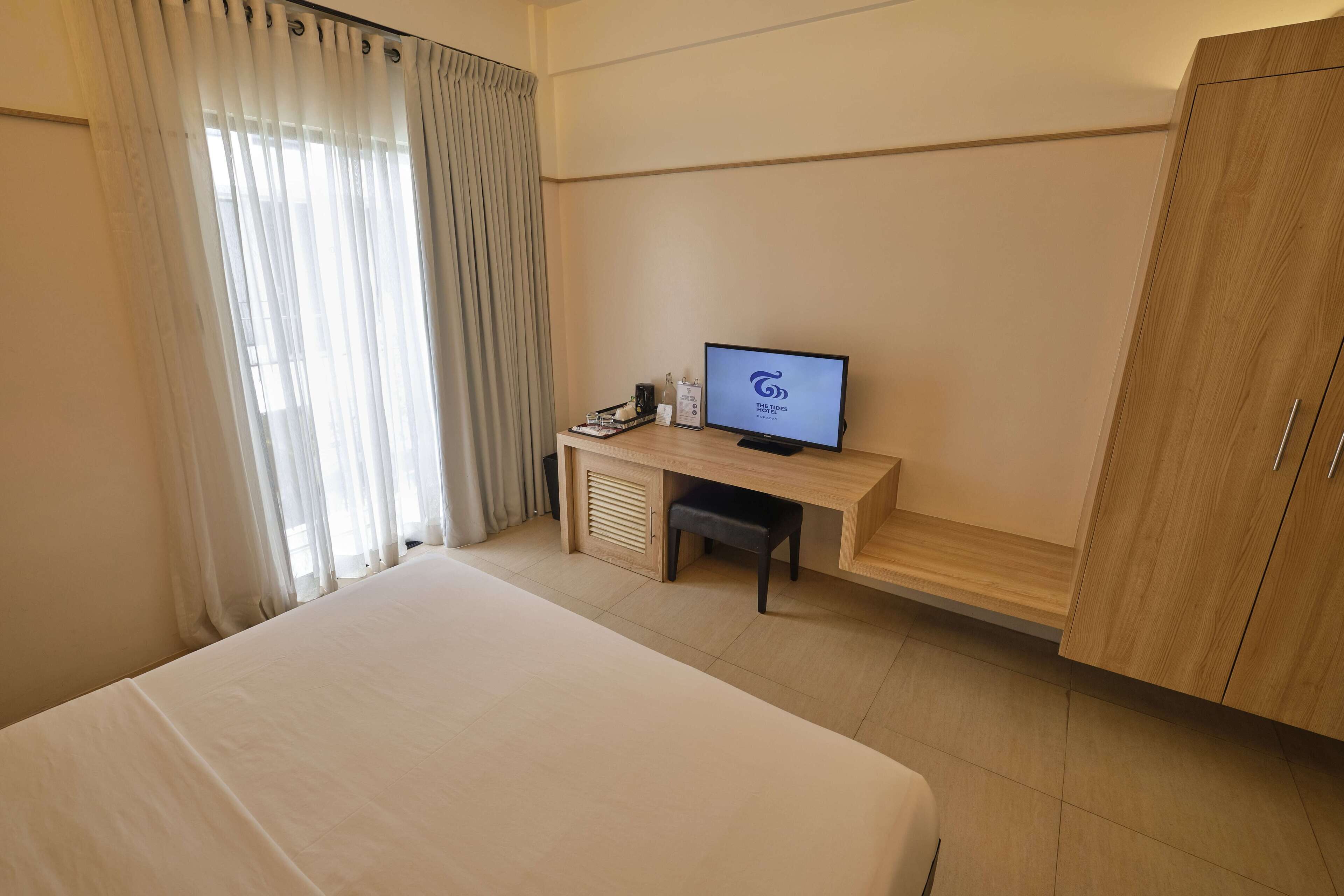 Habitación de hotel con una cama, un escritorio con computadora, un armario y una ventana con cortinas.