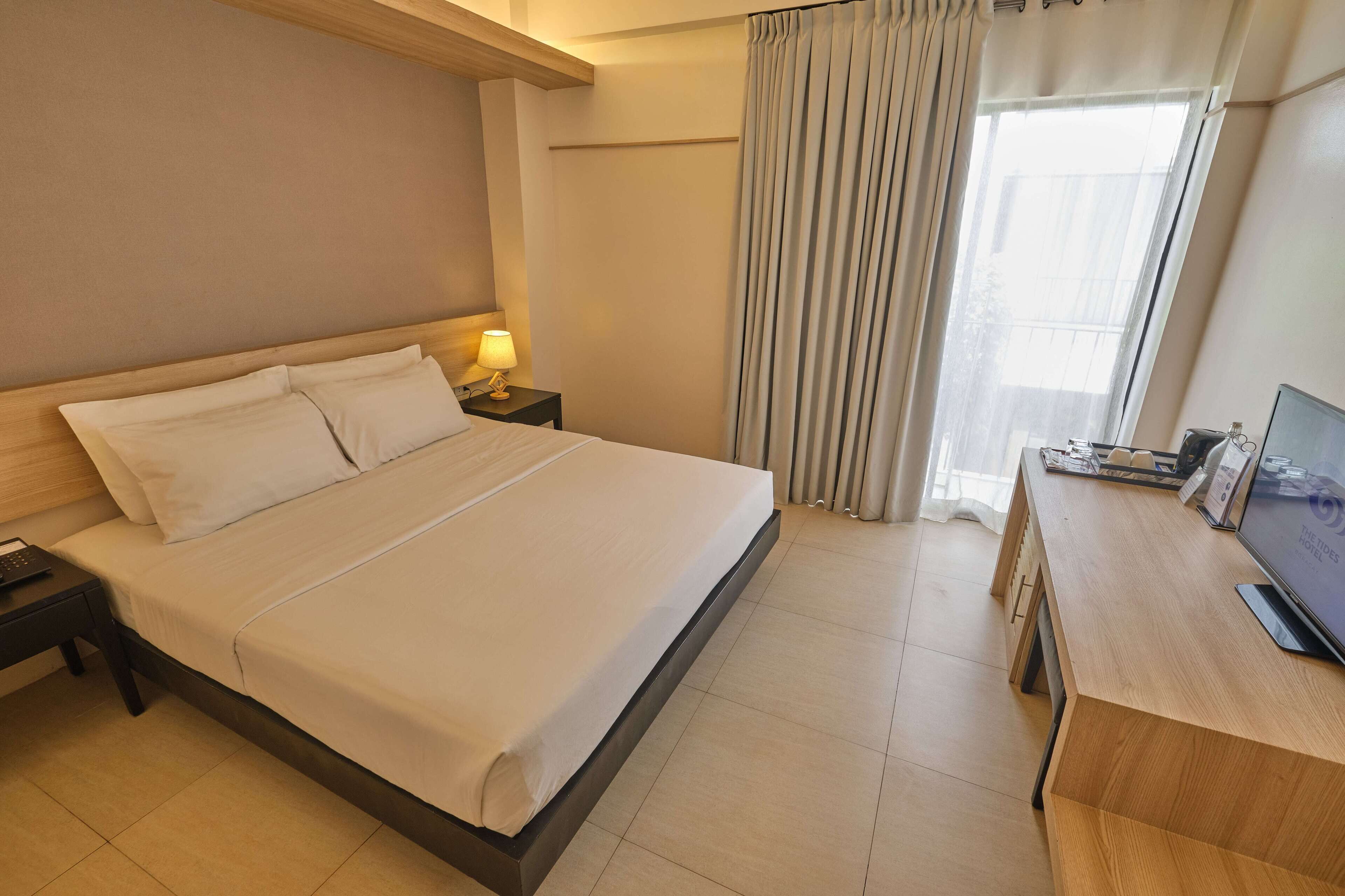 Une chambre d’hôtel avec un grand lit, une table de chevet avec une lampe, une télévision et une fenêtre avec des rideaux.