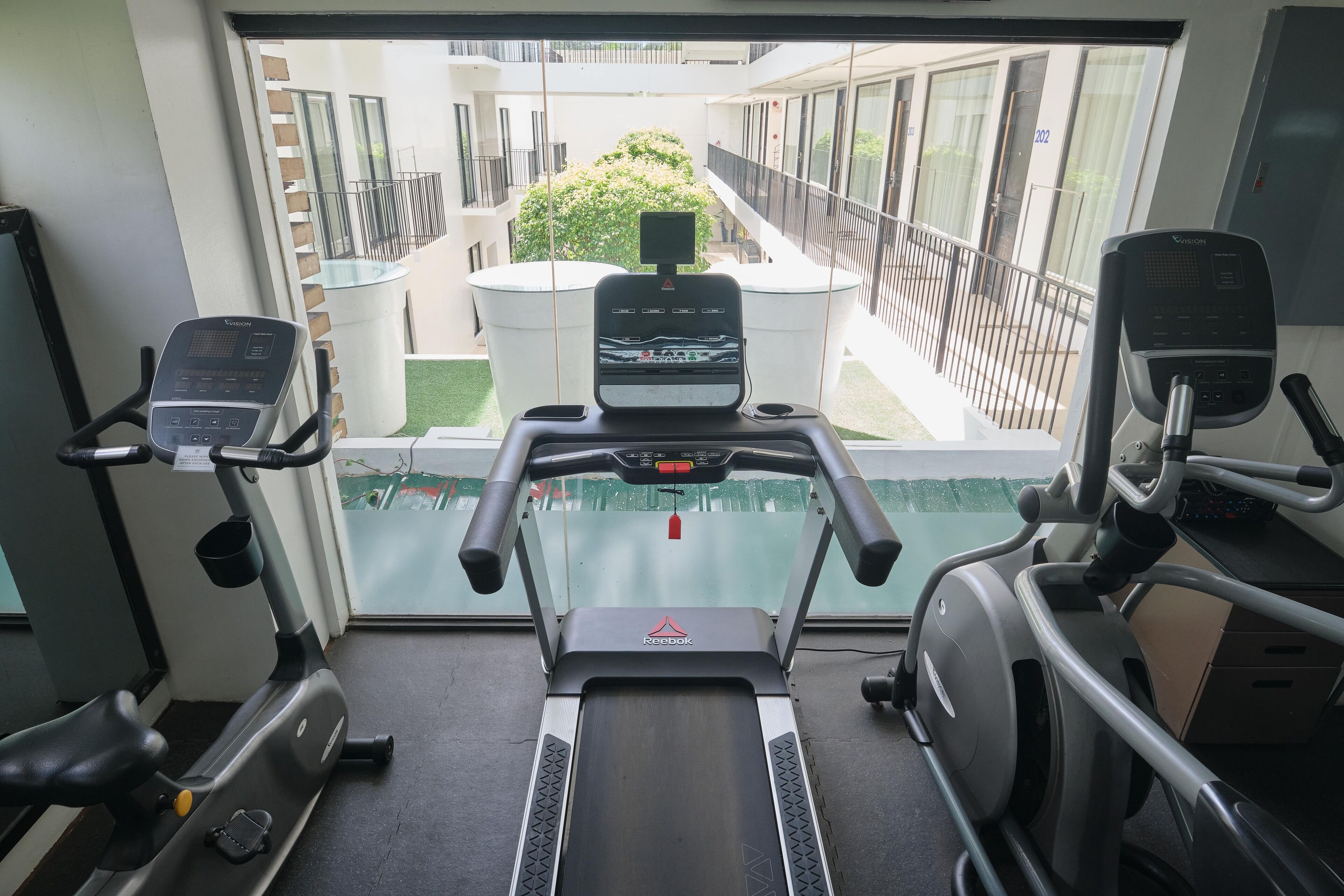 A gym with treadmills and exercise equipment, a window with a view of a building and a tree, and a door with a number.