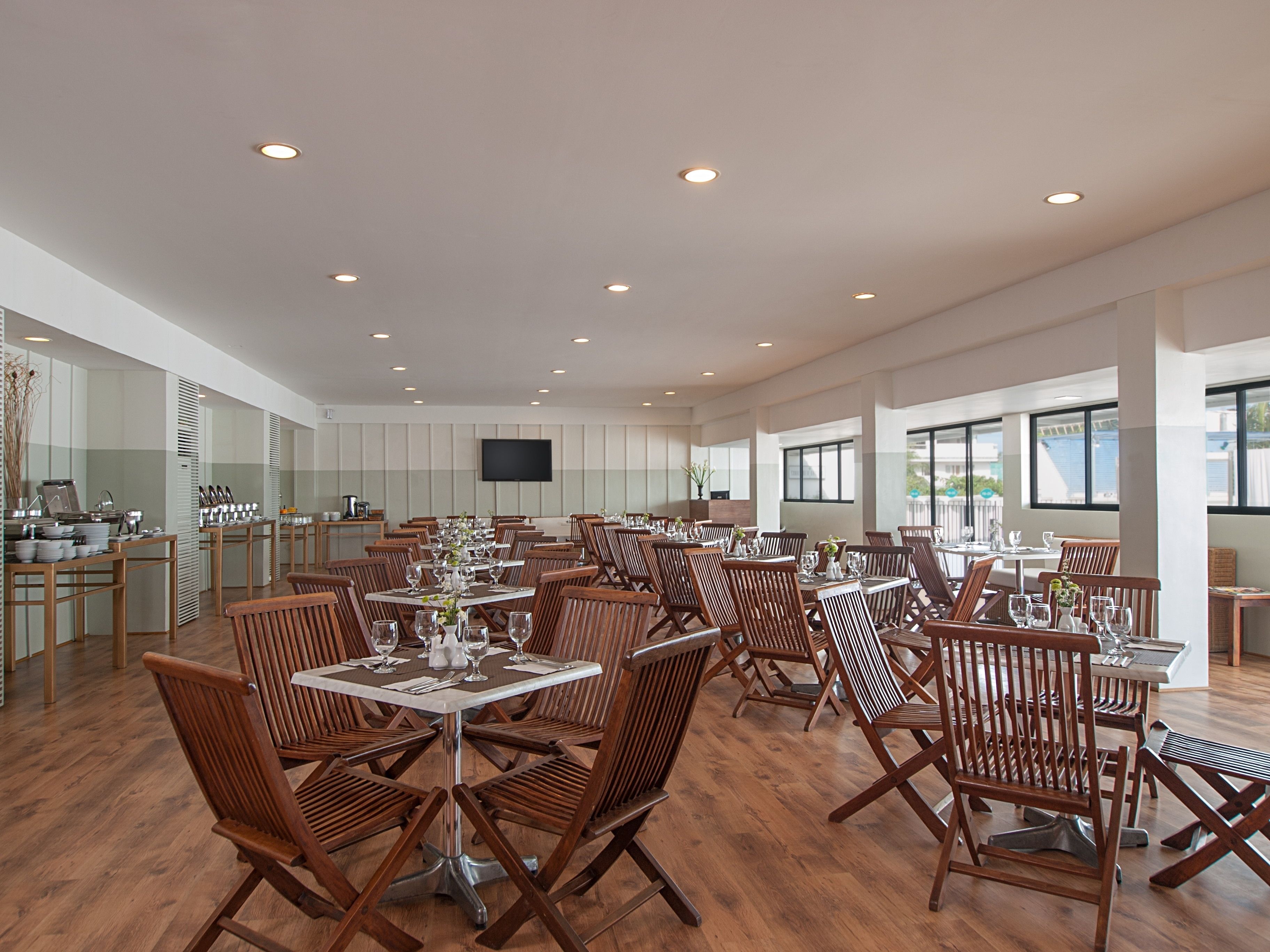 A spacious dining area with multiple tables and chairs, a flat-screen TV mounted on the wall, and large windows allowing natural light.