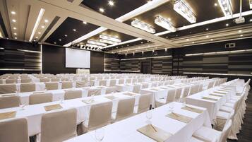 Un salón de conferencias con filas de mesas, cada una cubierta con un mantel blanco y posavasos dorados, equipado con copas de vino y cubiertos.