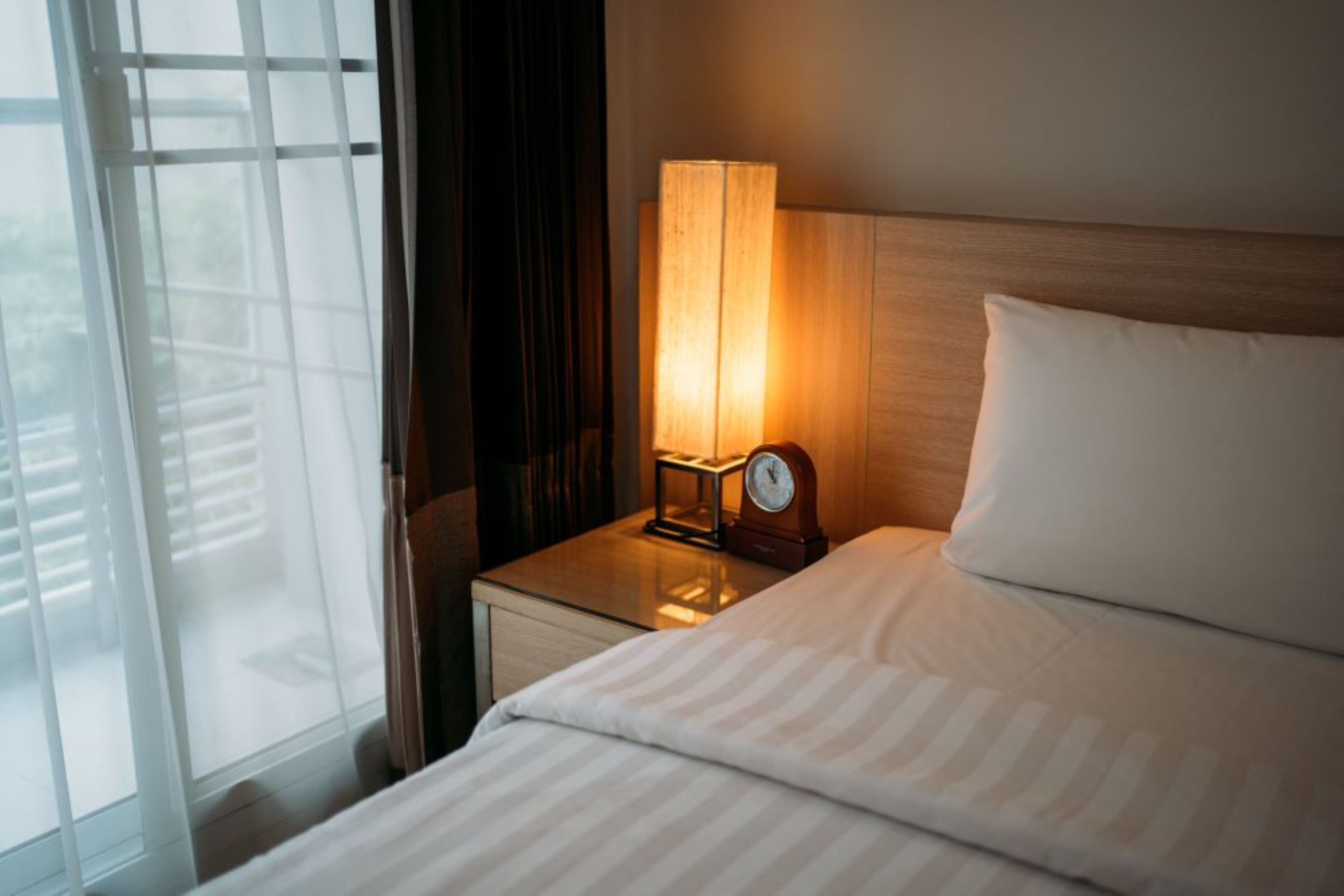A hotel room with a bed, a nightstand with a lamp, and a view through a window with blinds.