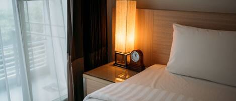 A hotel room with a bed, a nightstand with a lamp, and a view through a window with blinds.