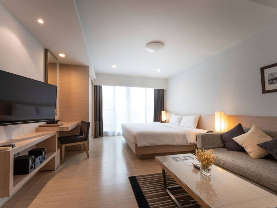 A modern hotel room with a large flat-screen TV, a bed with white linens, a sofa with cushions, a coffee table, and a desk with a computer and speakers.
