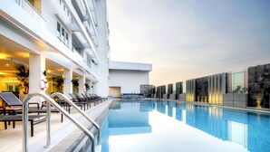 A swimming pool with sun loungers and a lifebuoy on the left, a modern building with balconies, and a clear sky.