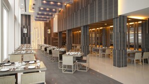 A modern dining area with multiple tables and chairs, large windows, and a high ceiling with recessed lighting.