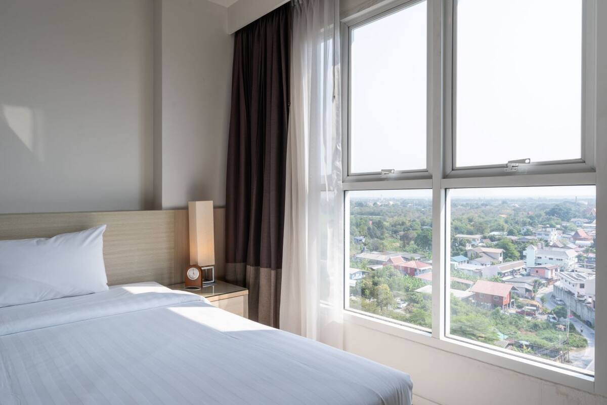 Habitación de hotel con una cama grande, una mesita de noche, un reloj y una ventana con vistas a la ciudad.