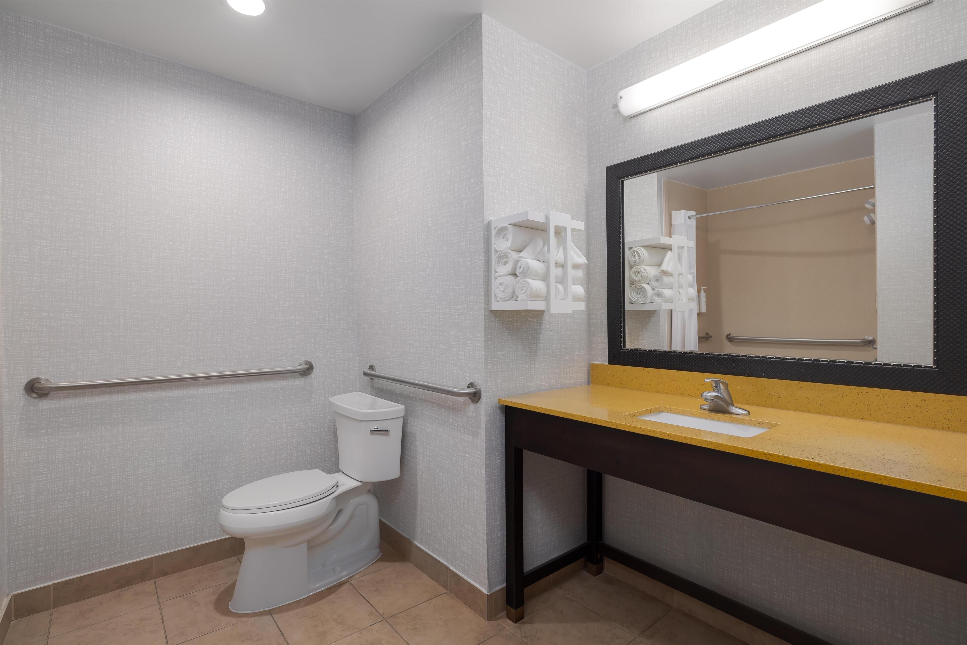 Un baño con inodoro, barras de apoyo, lavamanos con espejo y espacio para toallas.