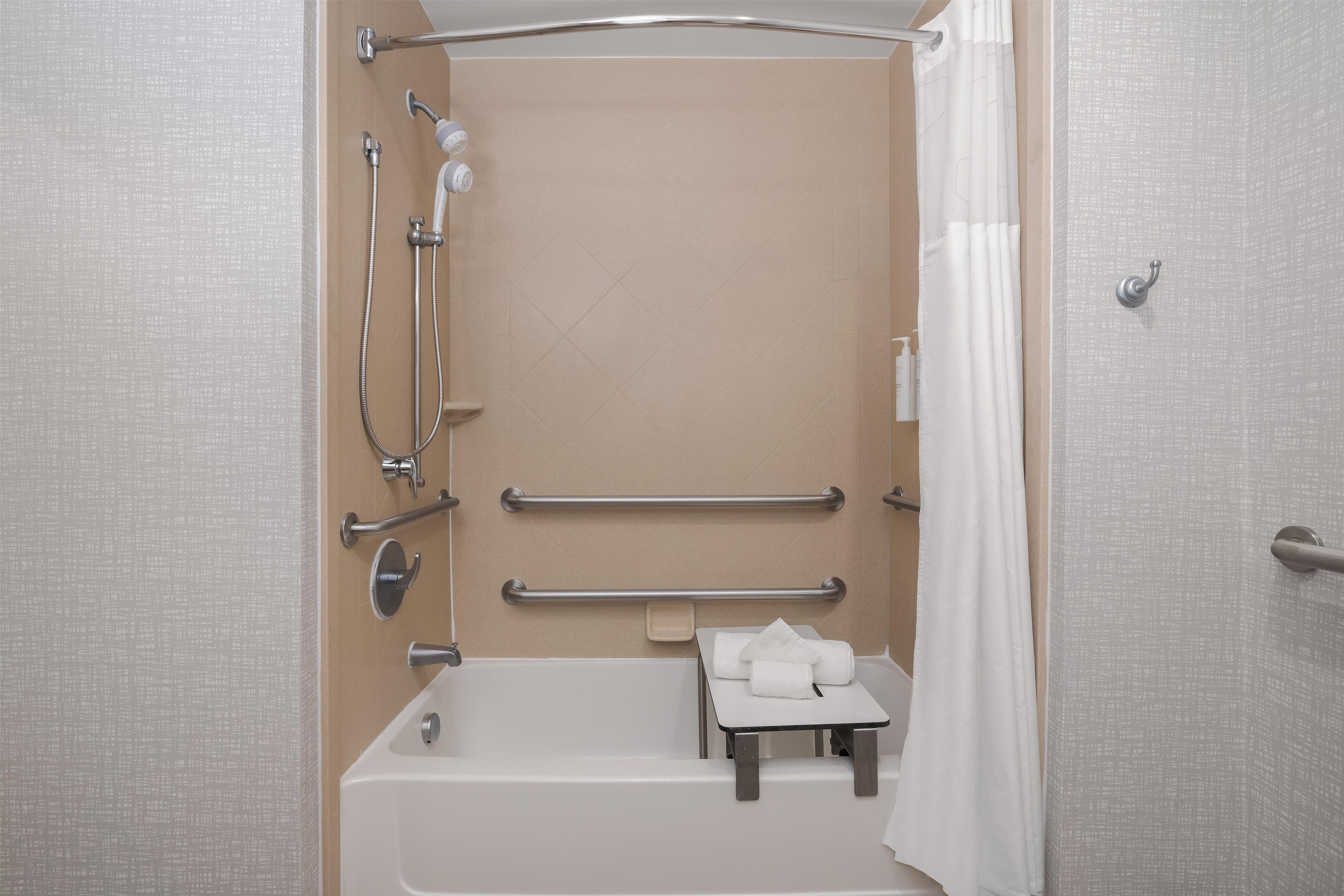 Une salle de bain équipée d’une baignoire, d’une douche, de barres d’appui, d’une petite table avec des serviettes et d’un rideau de douche blanc.