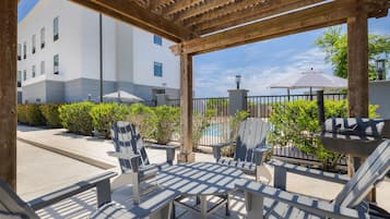 Un espacio de estar al aire libre cubierto con sillas Adirondack y una parrilla, una piscina al fondo y un edificio con fachada blanca.