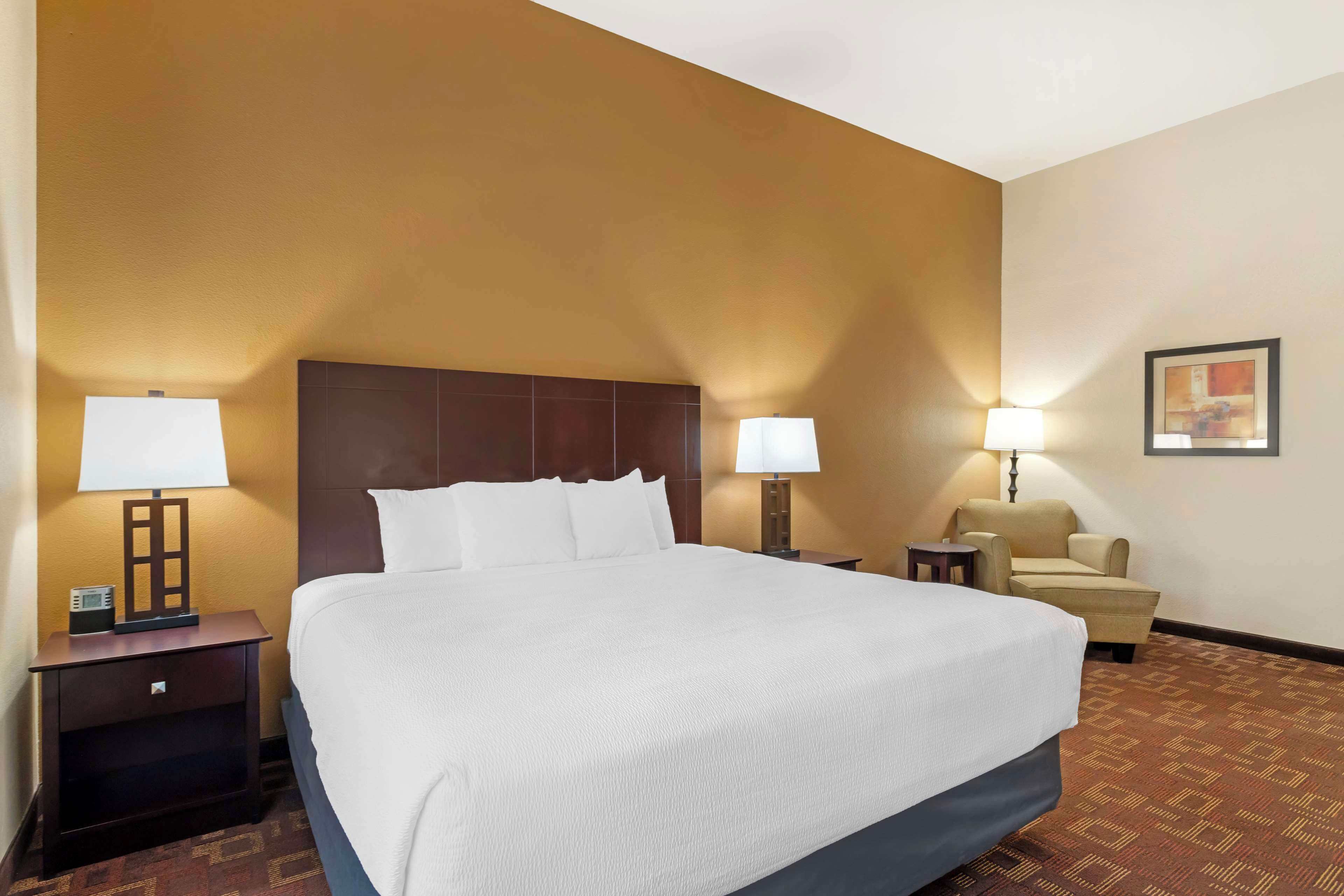 Una habitación de hotel con una cama grande, dos mesitas de noche con lámparas, una silla y un cuadro en la pared.