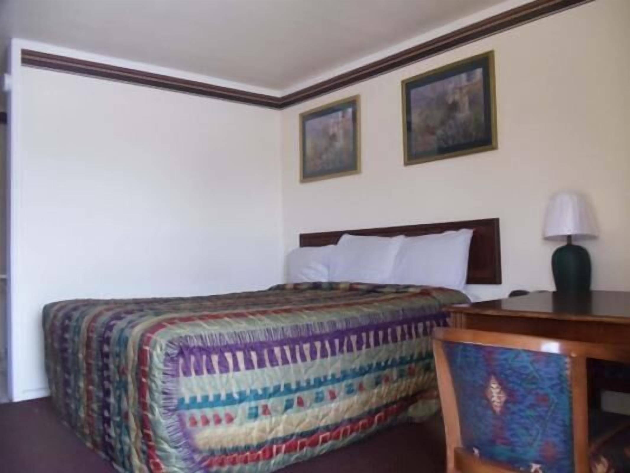 A hotel room with a neatly made bed, a wooden nightstand with a lamp, and two framed pictures on the wall.