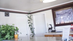 A reception area with a reception desk, a framed picture on the wall, a potted plant, and a thermostat on the wall. - Antlers Budget Inn (Antlers)