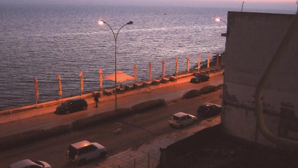 Una carretera costera con farolas, automóviles estacionados y vista al mar al anochecer.
