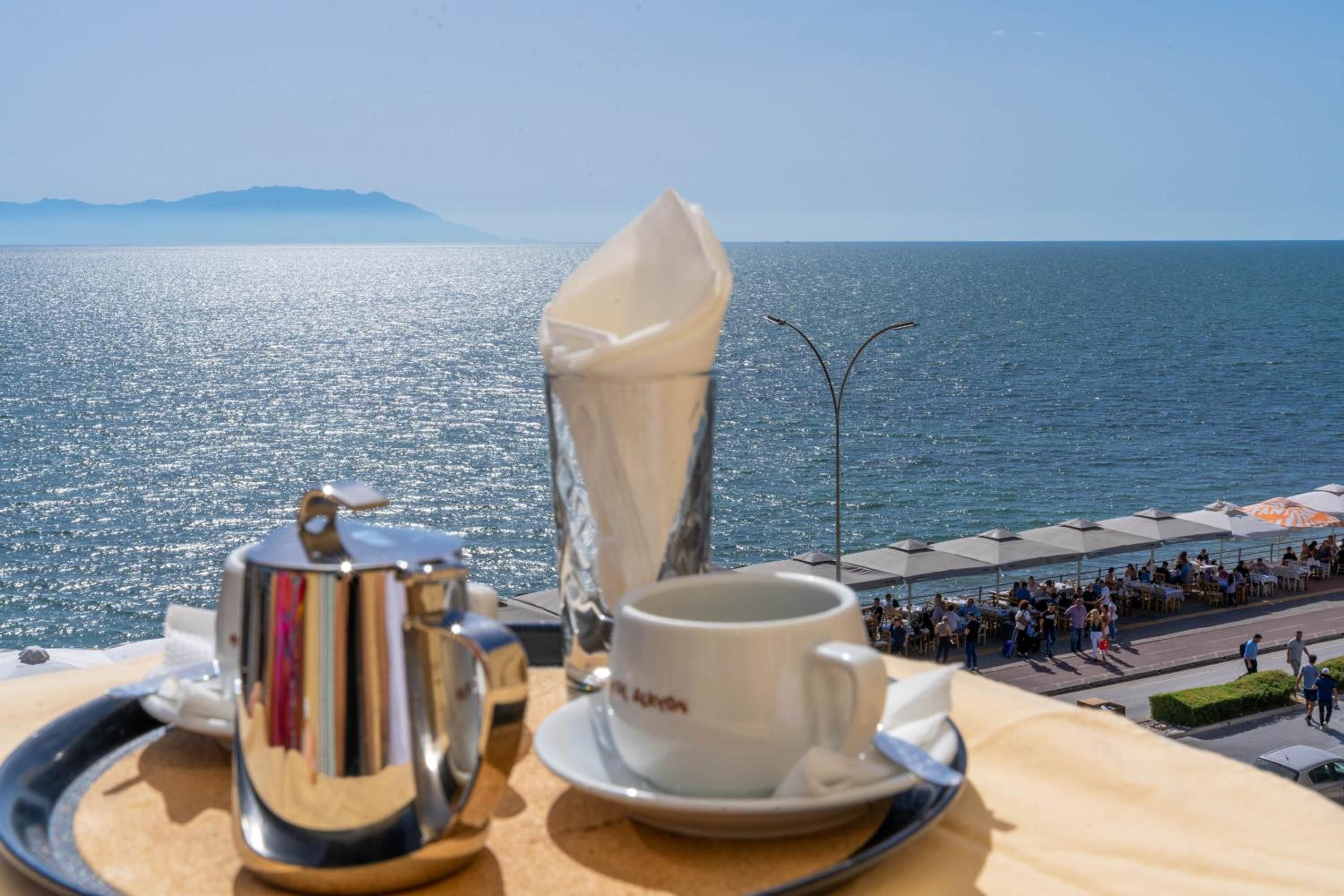 A table with a coffee pot, cup, and saucer, overlooking a coastal area with people and umbrellas.