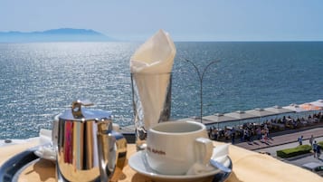 A table with a coffee pot, cup, and saucer, overlooking a coastal area with people and umbrellas.