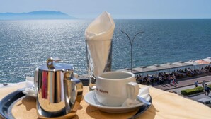 A table with a coffee pot, cup, and saucer, overlooking a coastal area with people and umbrellas. - Hotel Alkyon (Alexandroupoli)