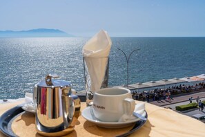 A table with a coffee pot, cup, and saucer, overlooking a coastal area with people and umbrellas. - Hotel Alkyon (Alexandroupoli)