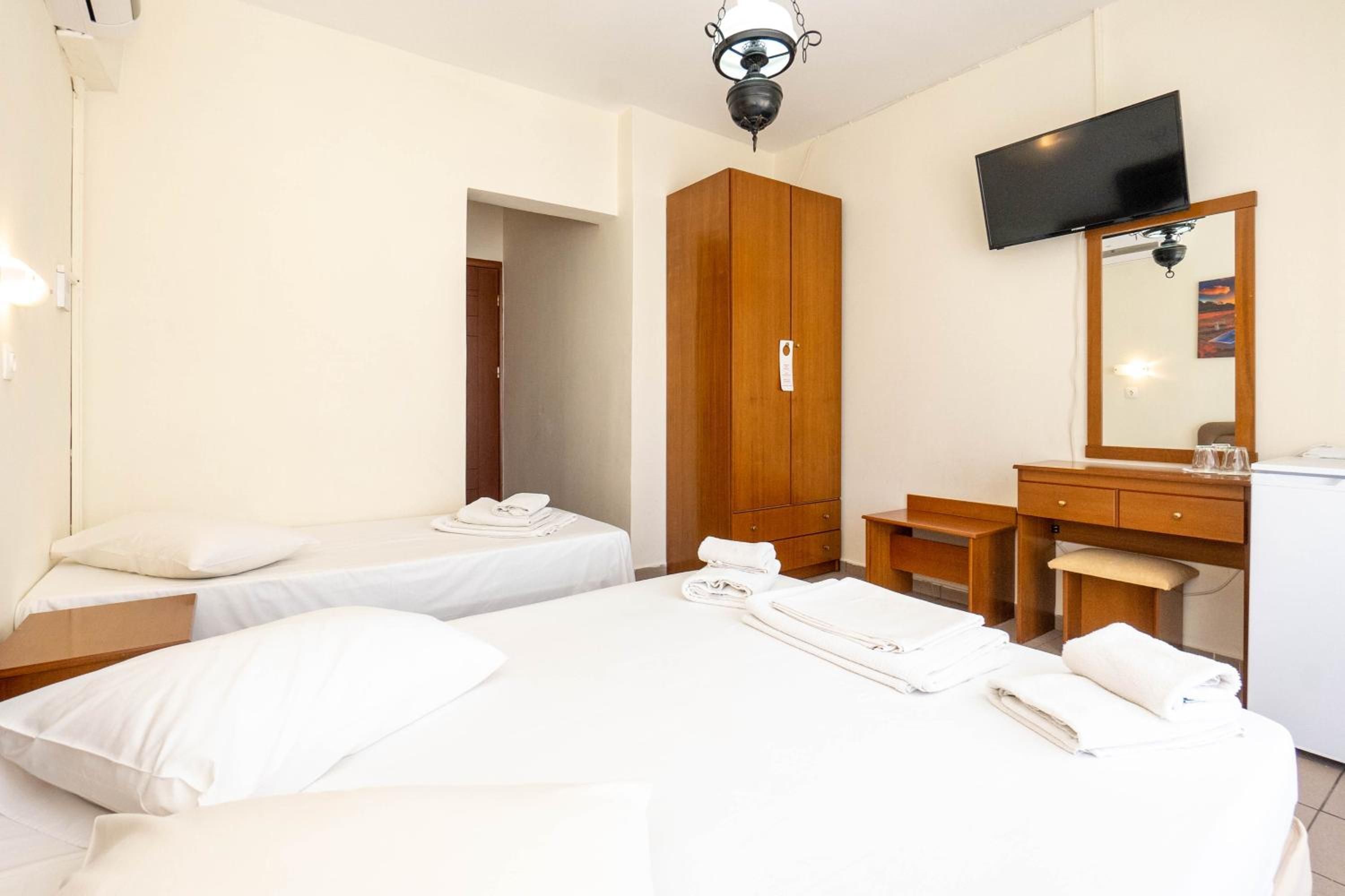 Habitación de hotel con dos camas, un armario de madera, un escritorio con silla, un televisor montado en la pared y una lámpara en el techo.