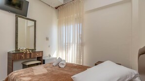 A hotel room with a bed, a wooden dresser, a mirror, a window with sheer curtains, and a flat-screen TV mounted on the wall.