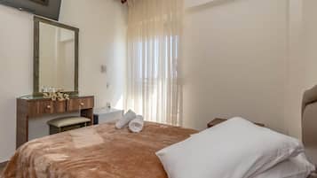 A hotel room with a bed, a wooden dresser, a mirror, a window with sheer curtains, and a flat-screen TV mounted on the wall.