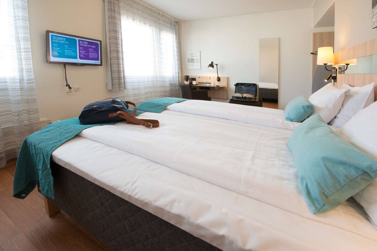 A hotel room with two beds, a desk, a chair, a TV, and a lamp.