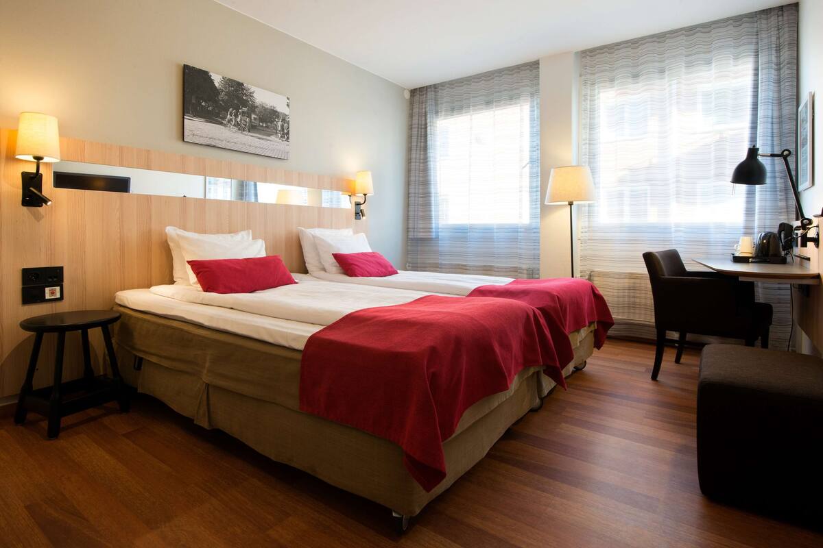 A hotel room with a double bed, two bedside tables, a desk with a chair, a lamp, and a large window with curtains.