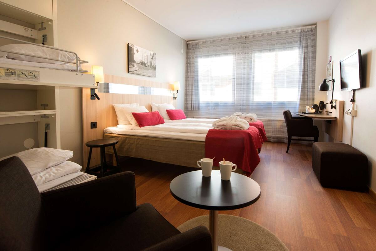 A hotel room with a bunk bed, a desk, a chair, a small round table, and a TV.