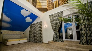 A room with a sloped ceiling, patterned curtains, a built-in bench, and a view of a cityscape through a glass door. - Mesogios Hotel (Chisinau)