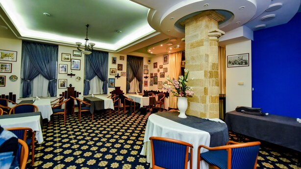 A restaurant with a patterned floor, tables with white tablecloths, blue chairs, and a central column with a vase of flowers. - Mesogios Hotel (Chisinau)