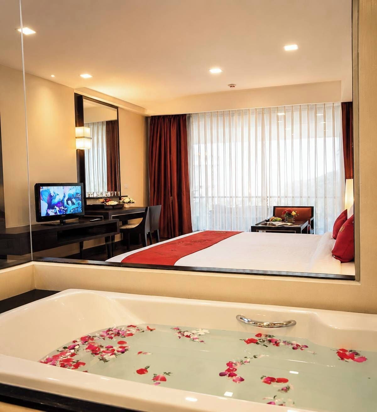 A hotel room with a large bed, a TV, a desk, a chair, a bathtub with floral decorations, and a large window with curtains.