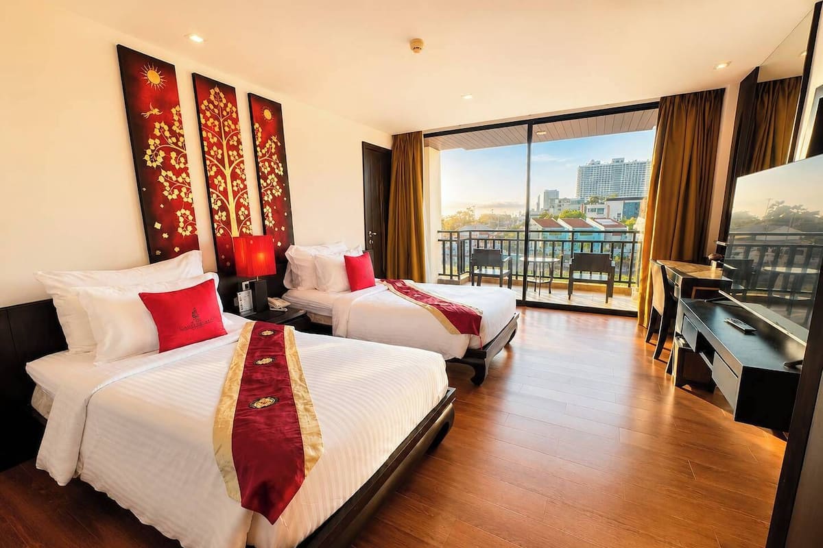 A hotel room with two beds, a desk, a TV, and a balcony with a city view.