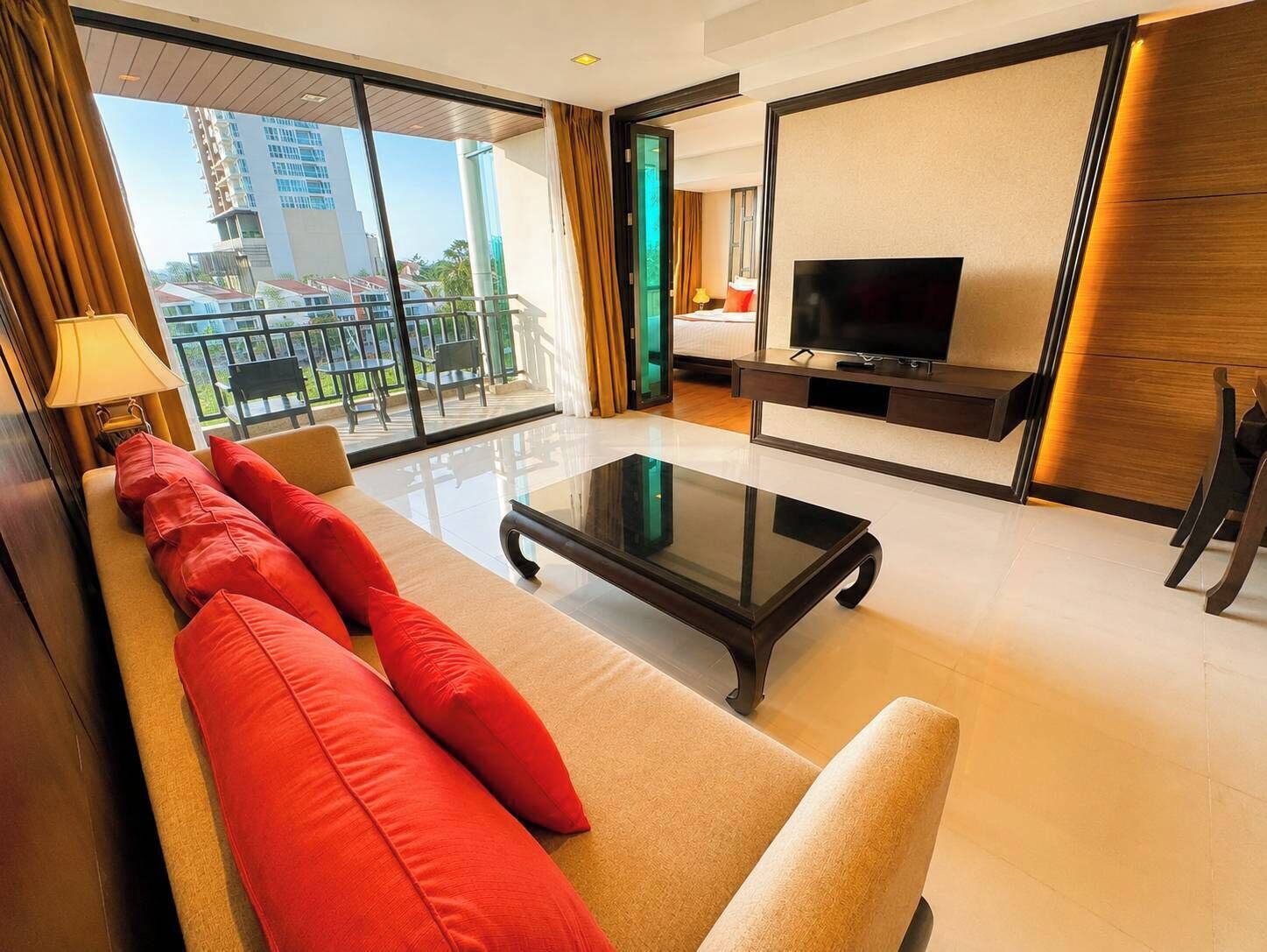 A modern hotel room with a sofa, a glass coffee table, a flat-screen TV, and a balcony with a view of buildings.