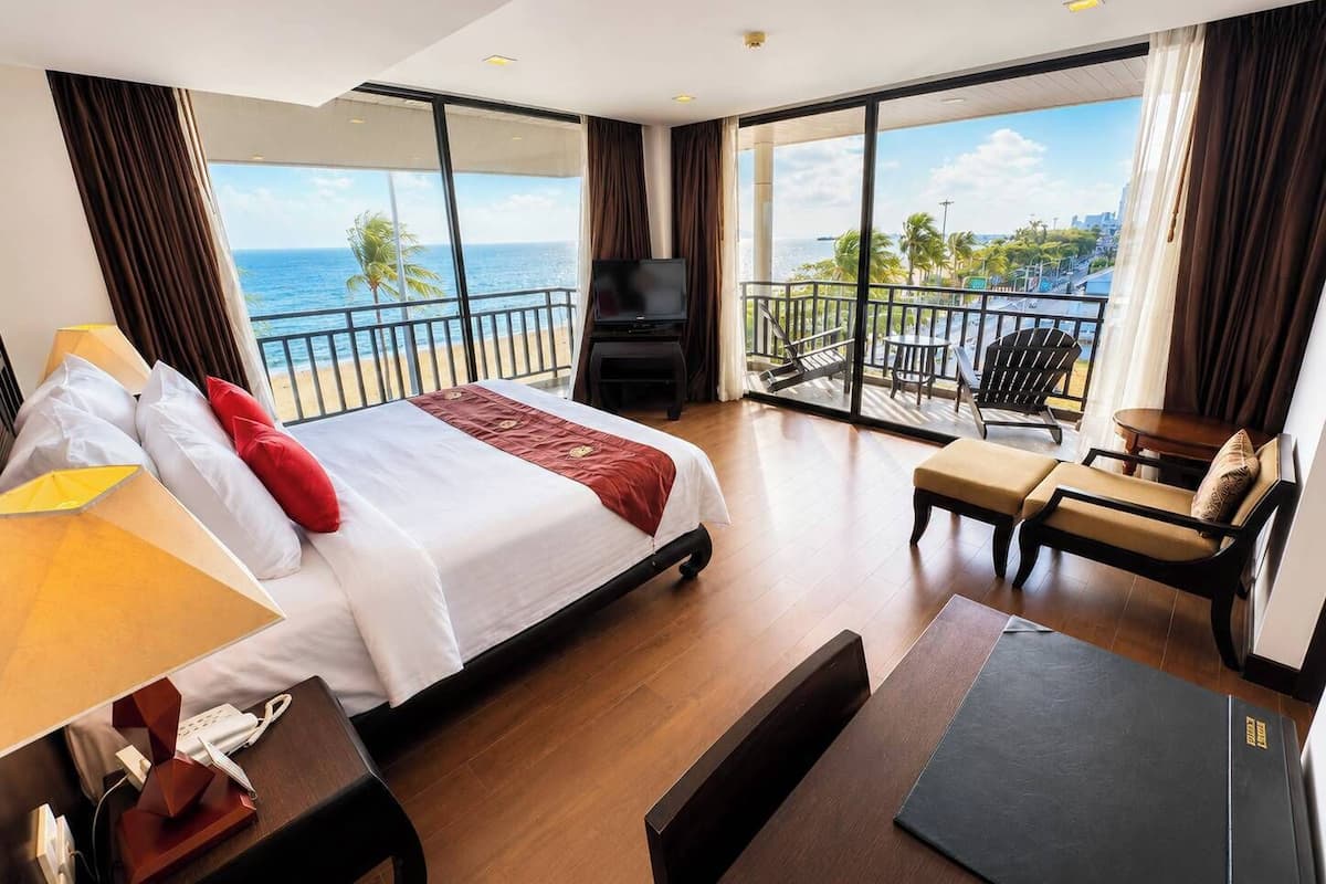 A hotel room with a large bed, a TV, a balcony with a view of the ocean, and a seating area with a table and chairs.