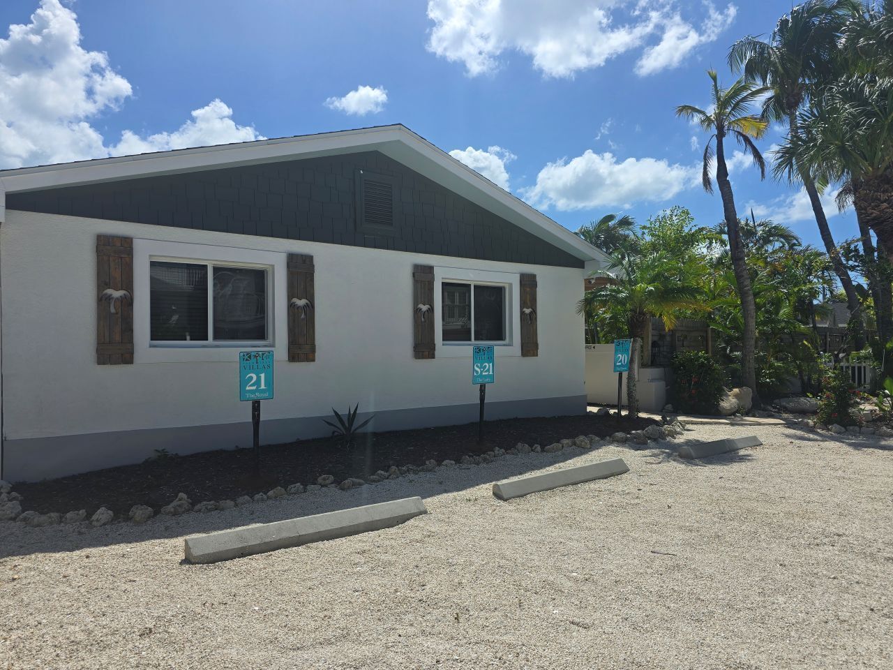 A single-story building with two windows, each with wooden shutters. There are two street signs in front, one labeled '21' and the other '20'. The building is surrounded by a gravel driveway and has palm trees in the background.