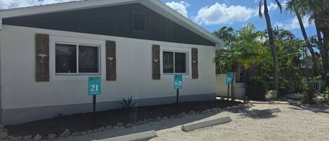 A single-story building with two windows, each with wooden shutters. There are two street signs in front, one labeled '21' and the other '20'. The building is surrounded by a gravel driveway and has palm trees in the background.