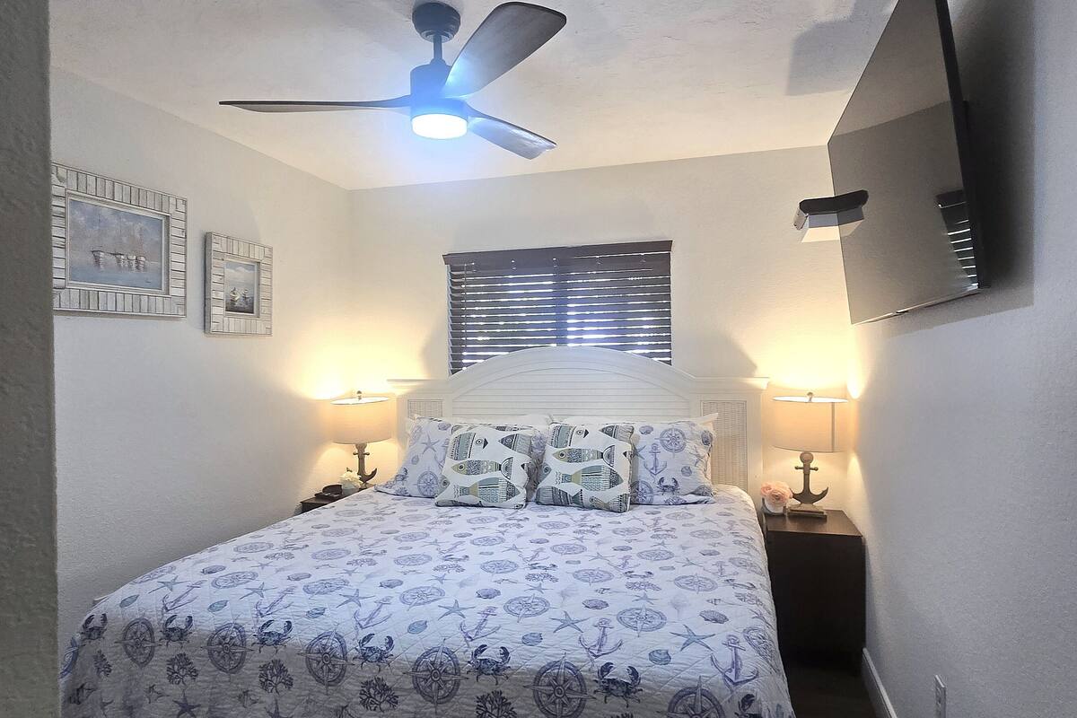 A bedroom with a bed, two bedside lamps, a ceiling fan, a window with blinds, and framed pictures on the wall.