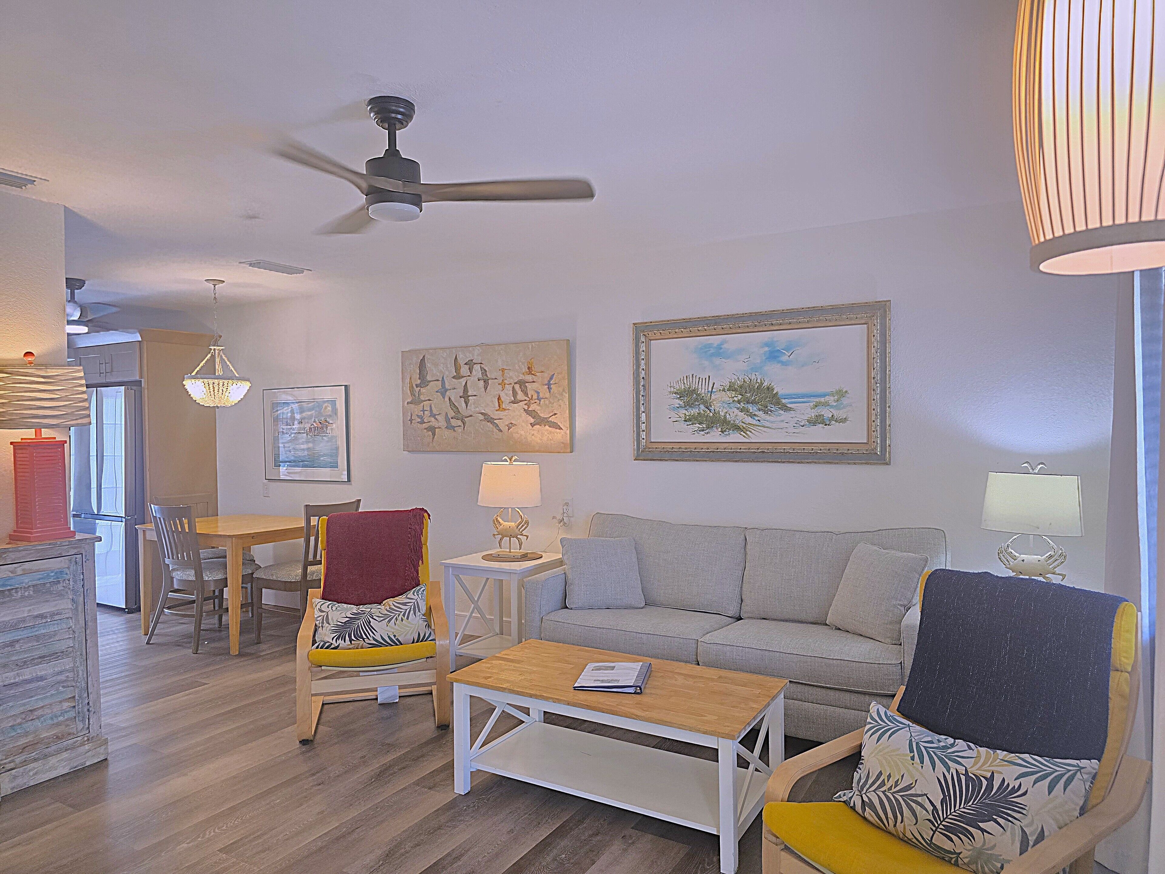 Un salon comprenant un canapé, une table basse, une table à manger avec des chaises, un ventilateur de plafond et des œuvres d’art accrochées aux murs.