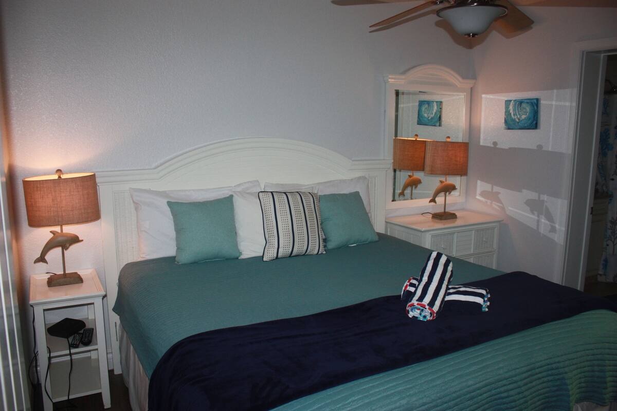 A neatly made bed with a teal bedspread and pillows, a bedside lamp, a nightstand with a remote control, and a ceiling fan.