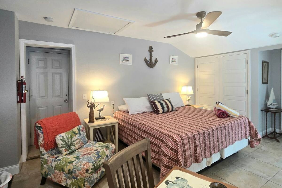 A bedroom with a bed, a chair, a table, a ceiling fan, and a door.